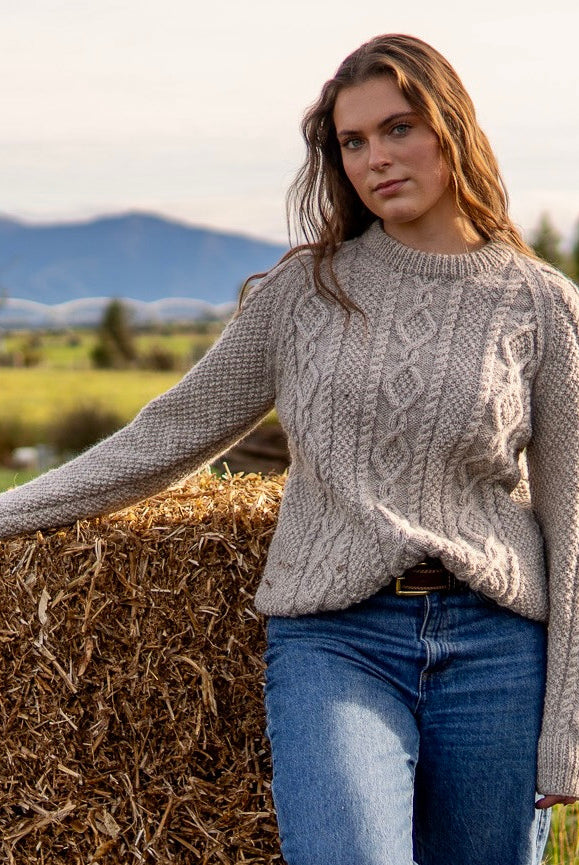 Woman leaning on hay wearing Lesley Unisex Aran Wool sweater in the colour Oatmeal.