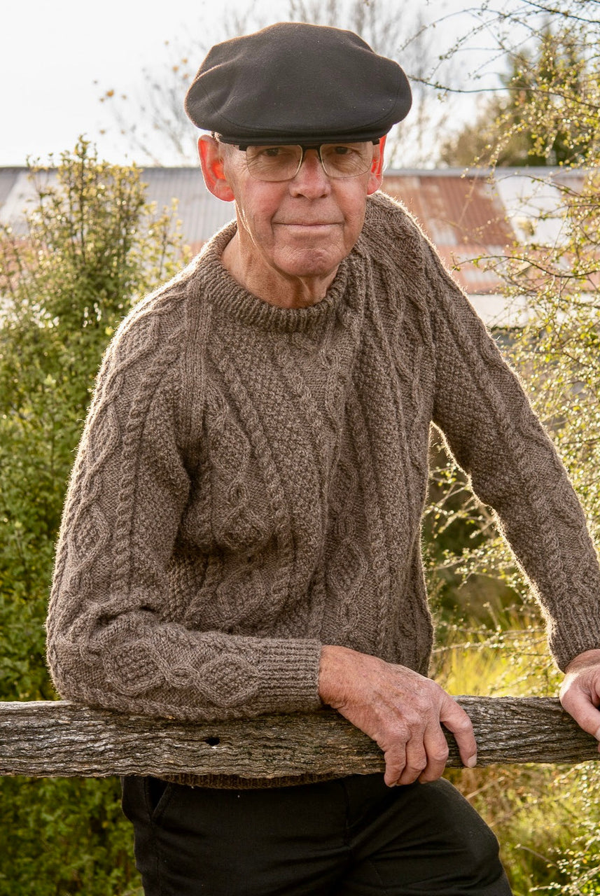 Man leaning on fence wearing Lesley Unisex Aran Wool sweater in the colour Brown.