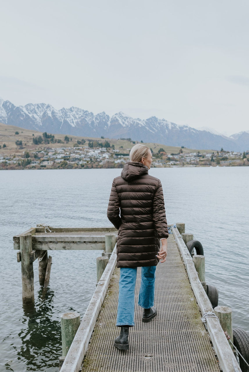 Woman wearing Moke - Arnie Reversible Packable Down Coat (ARNIE) in colour Chocolate/Palomino walking on lake pier.