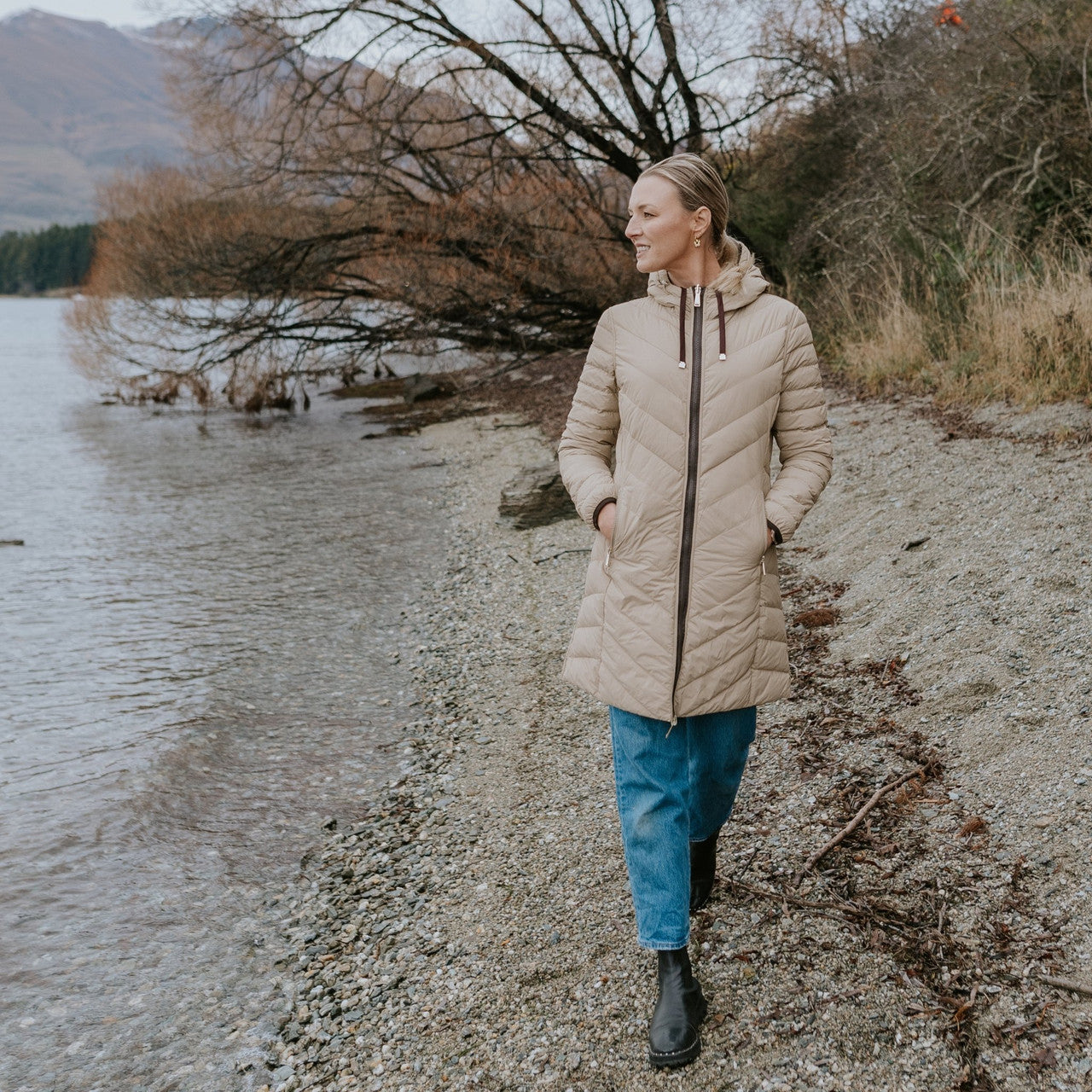 Woman wearing Moke - Arnie Reversible Packable Down Coat (ARNIE) in colour Chocolate/Palomino walking on lakefront.