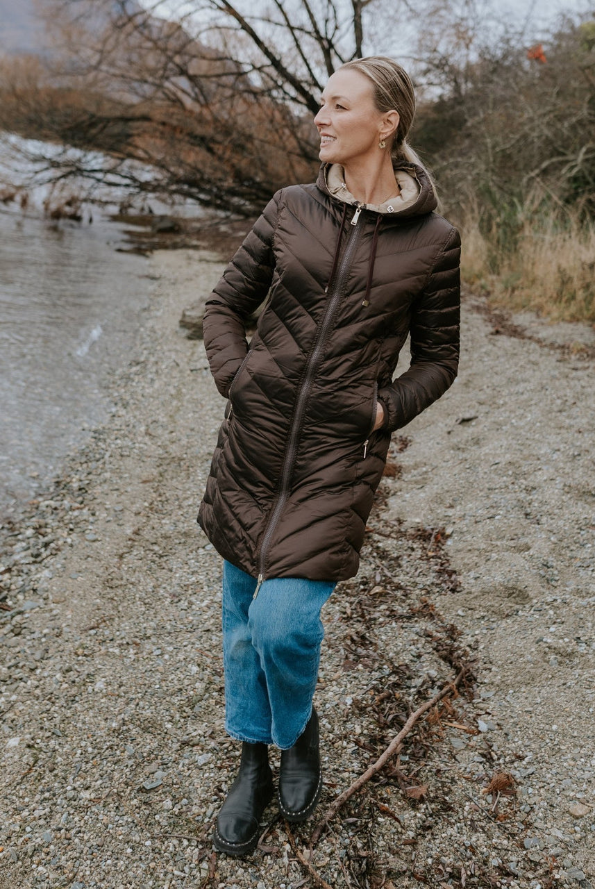 Woman wearing Moke - Arnie Reversible Packable Down Coat (ARNIE) in colour Chocolate/Palomino walking on lakefront.