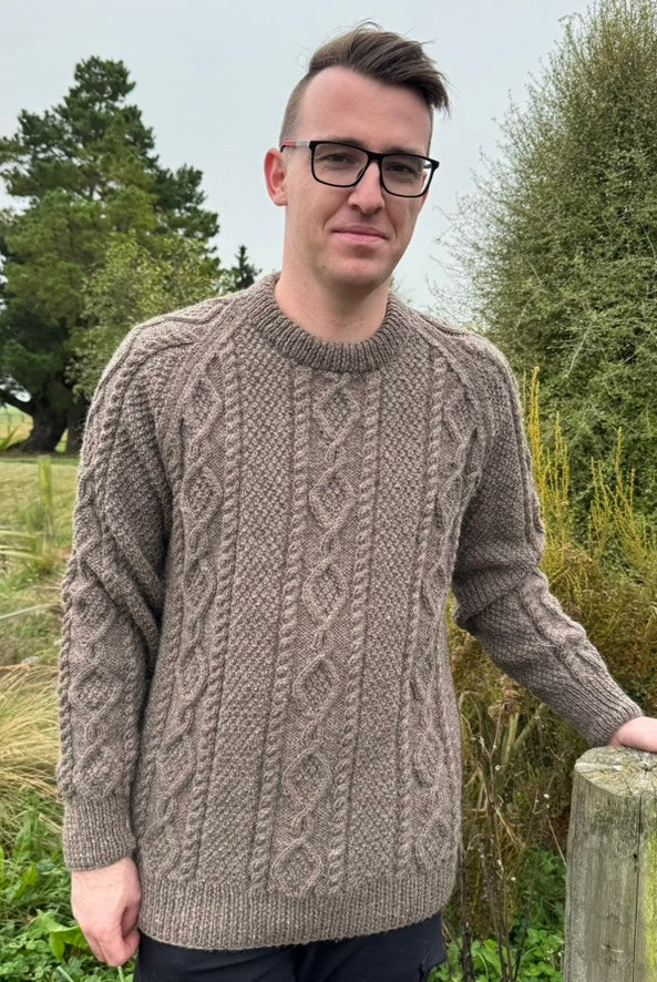 Man leaning on fence wearing Lesley Unisex Aran Wool sweater in the colour Brown.