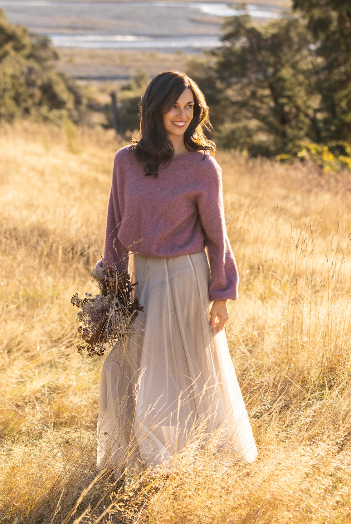 A woman stands in a sunlit field of dry golden grass, wearing a dusty mauve knit sweater and a long cream tulle skirt, holding a small bouquet of dried flowers.