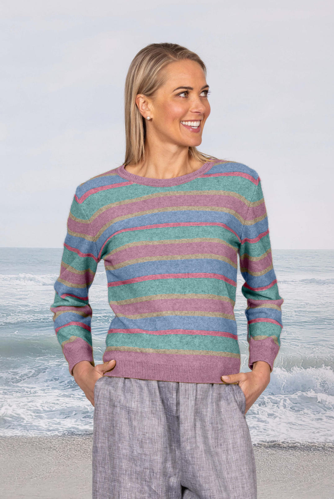A woman stands on a beach wearing a long-sleeve knit sweater with horizontal stripes in teal, pink, and lavender, paired with light gray pants.