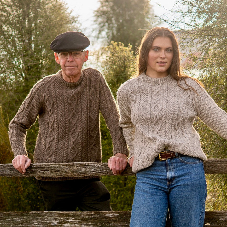 Man leaning on fence wearing Lesley Unisex Aran Wool sweater in the colour Brown and woman wearing the colour Oatmeal.