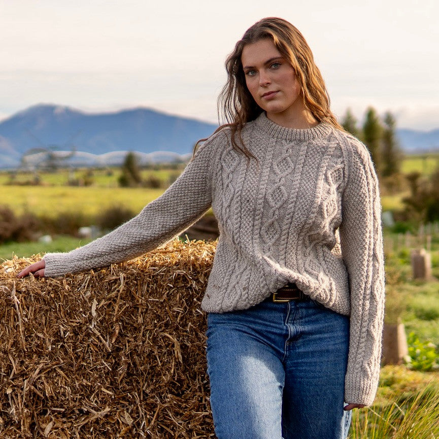 Woman leaning on hay wearing Lesley Unisex Aran Wool sweater in the colour Oatmeal.