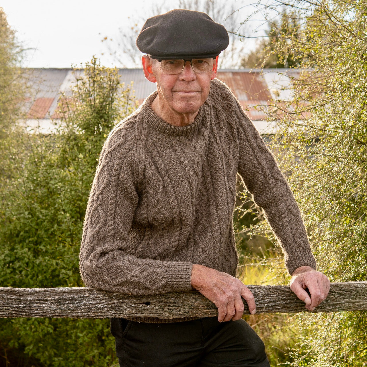 Man leaning on fence wearing Lesley Unisex Aran Wool sweater in the colour Brown.