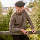 Man leaning on fence wearing Lesley Unisex Aran Wool sweater in the colour Brown.
