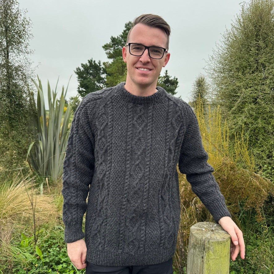 Man leaning on fence wearing Lesley Unisex Aran Wool sweater in the colour Charcoal.