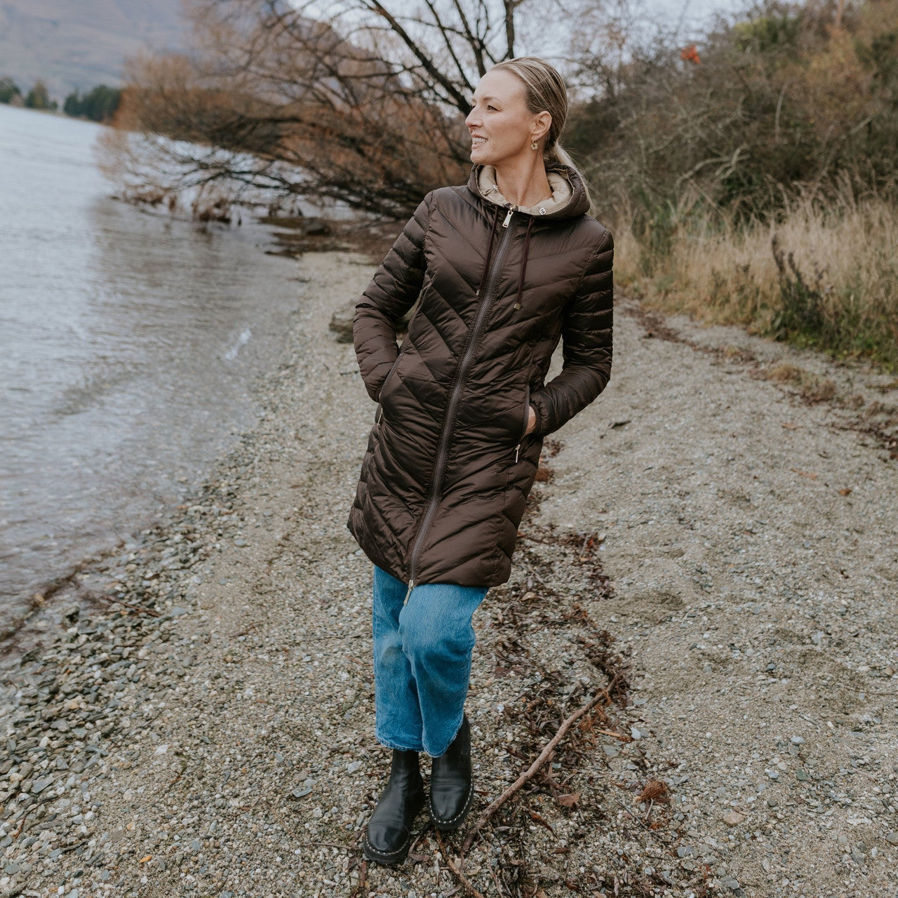 Woman wearing Moke - Arnie Reversible Packable Down Coat (ARNIE) in colour Chocolate/Palomino walking on lakefront.