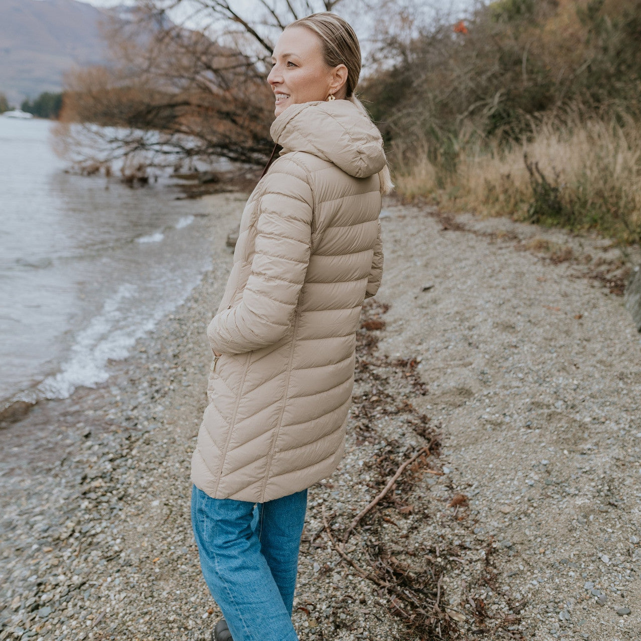 Woman wearing Moke - Arnie Reversible Packable Down Coat (ARNIE) in colour Chocolate/Palomino walking on lakefront.