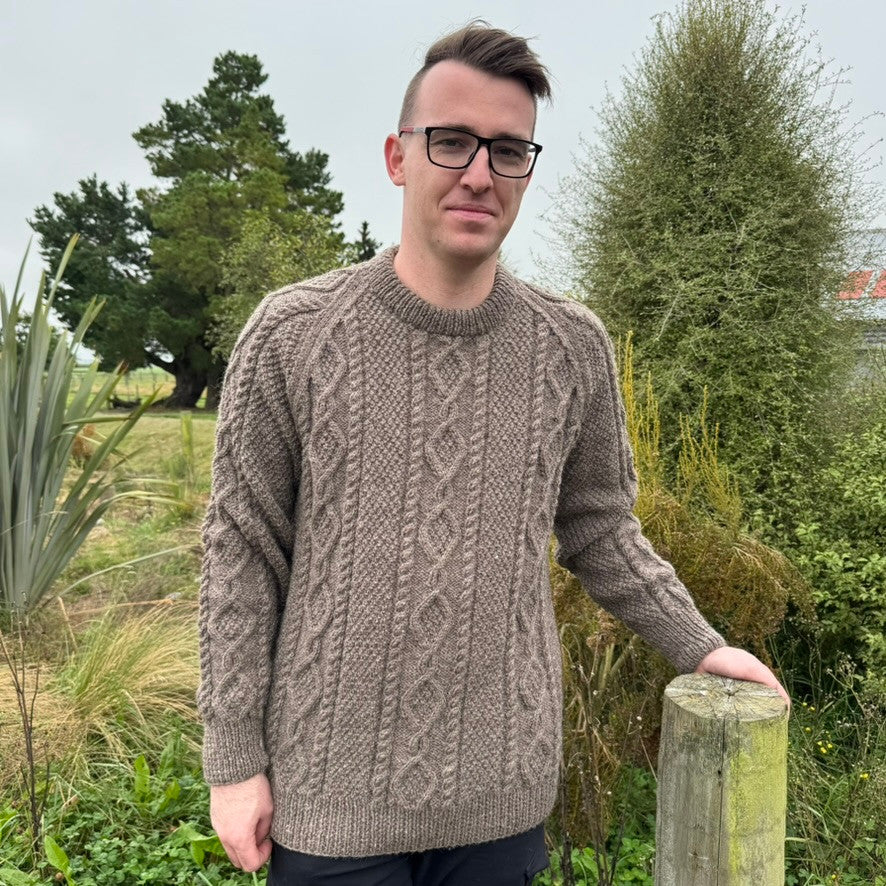 Man leaning on fence wearing Lesley Unisex Aran Wool sweater in the colour Brown.