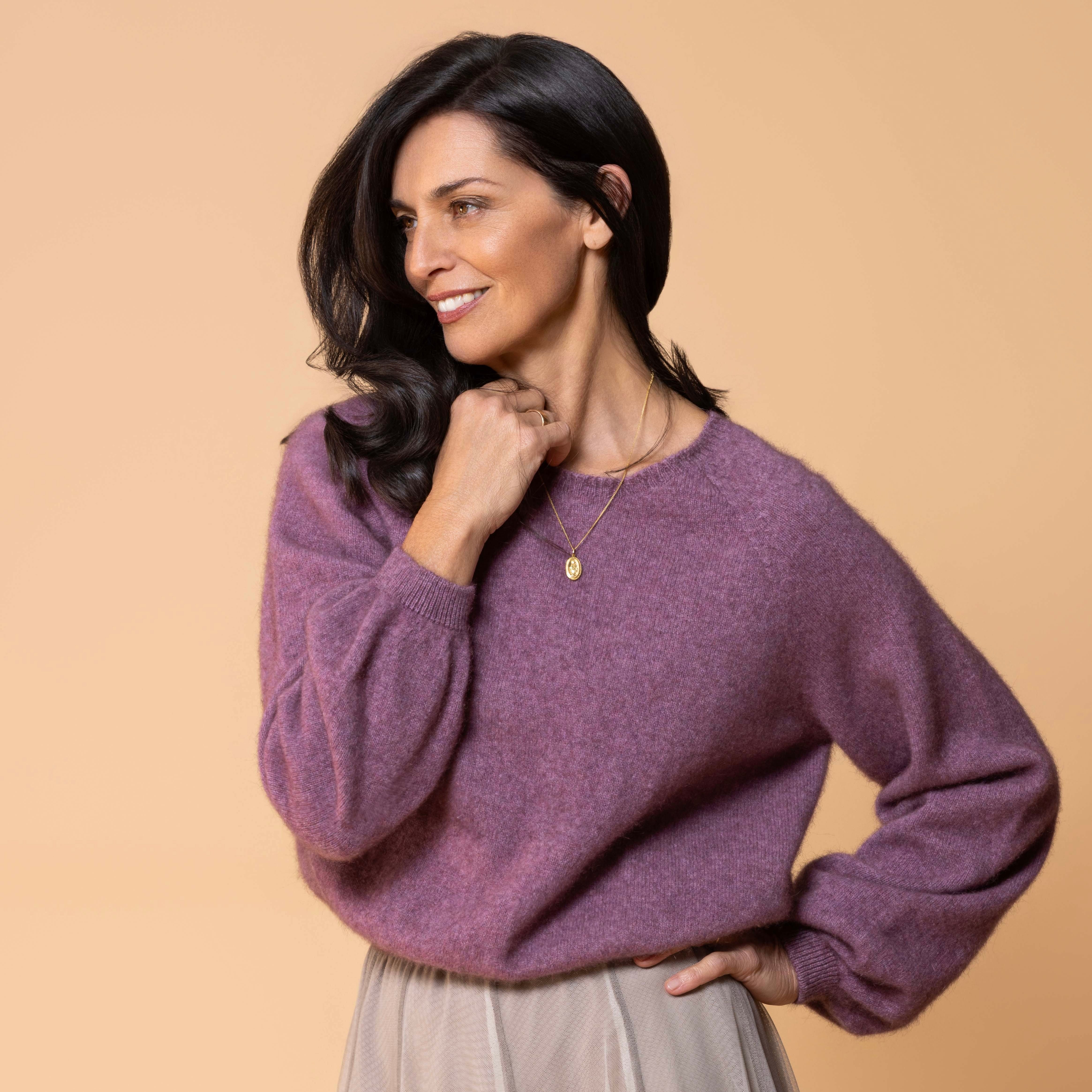 A woman wearing a heathered purple knit sweater and a pleated light beige skirt, posing against a solid beige background.