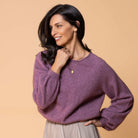 A woman wearing a heathered purple knit sweater and a pleated light beige skirt, posing against a solid beige background.