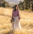 A woman stands in a sunlit field of dry golden grass, wearing a dusty mauve knit sweater and a long cream tulle skirt, holding a small bouquet of dried flowers.