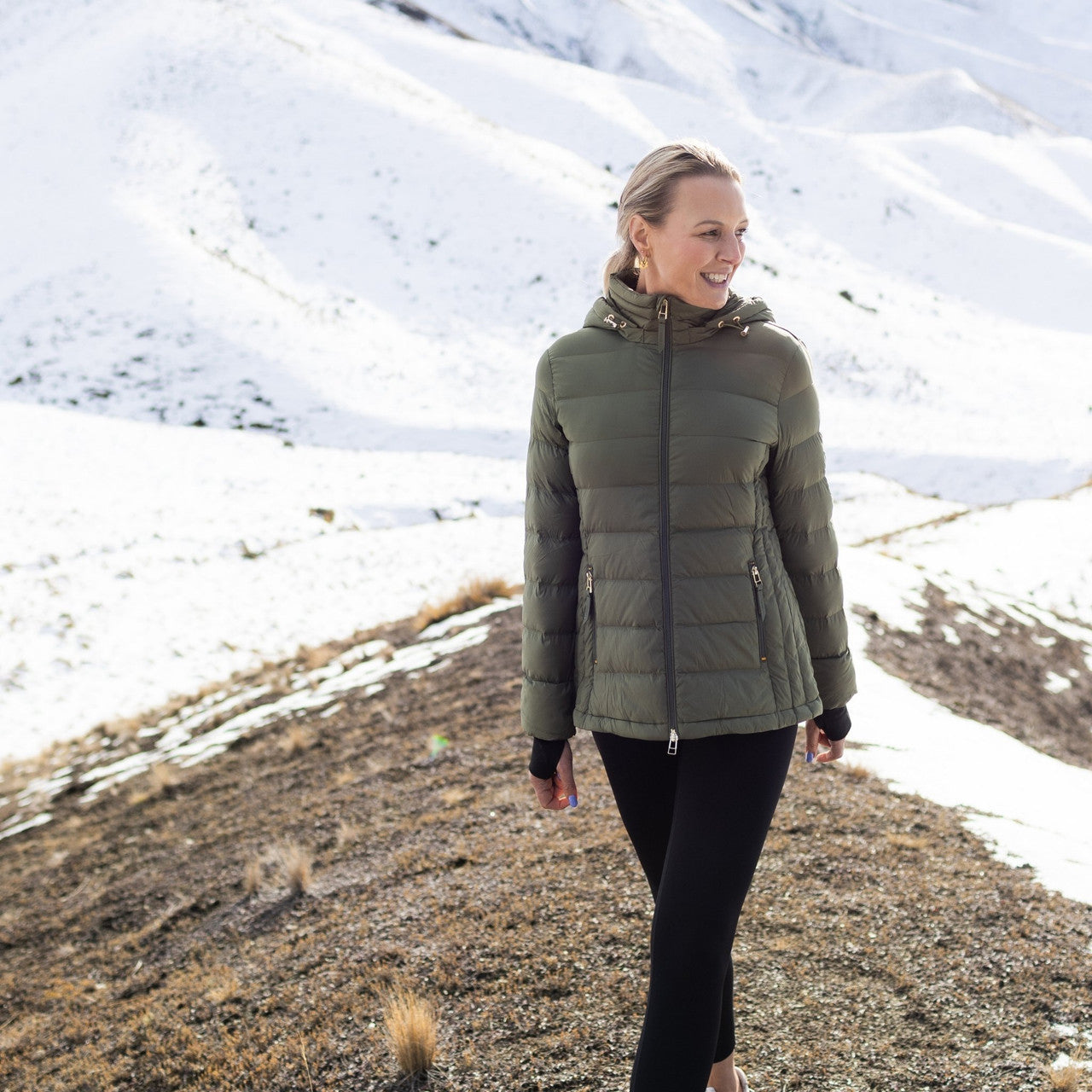 Woman wearing Moke - Lucy Cloud Touch Packable Down Jacket (LUCY) in the colour Olive Green on a hill with snow in the background.