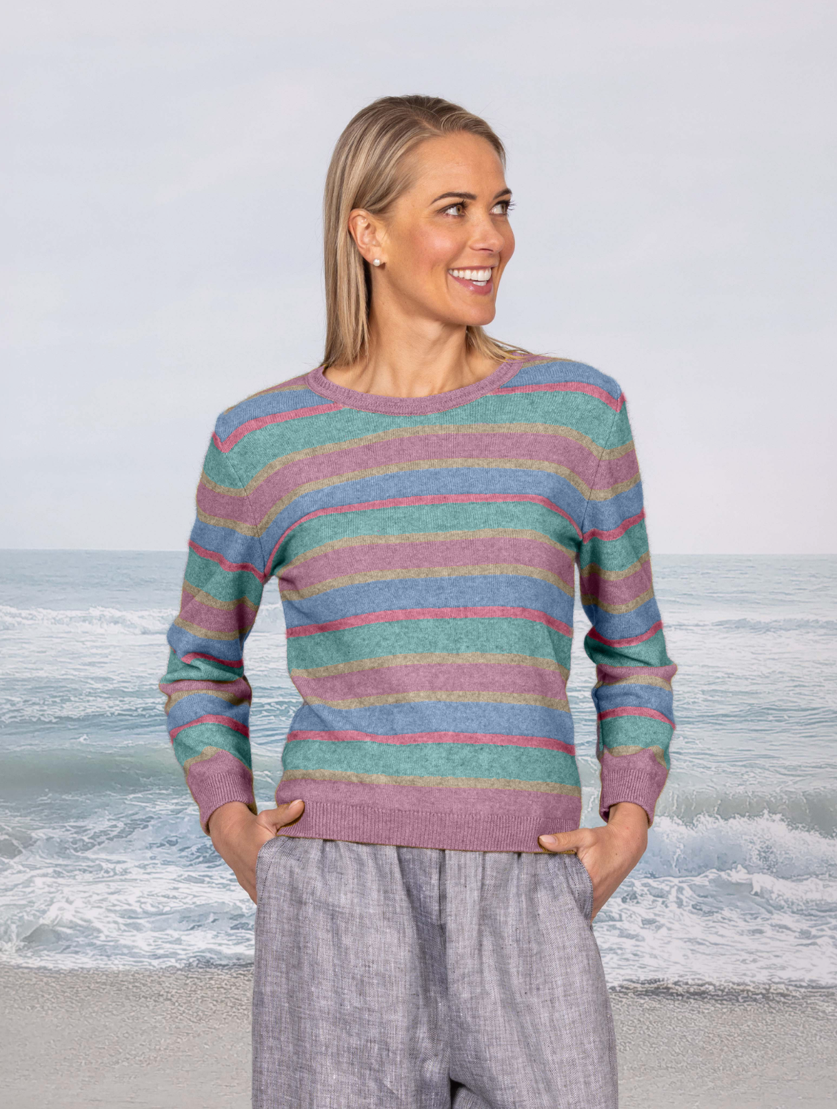 A woman stands on a beach wearing a long-sleeve knit sweater with horizontal stripes in teal, pink, and lavender, paired with light gray pants.