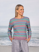 A woman stands on a beach wearing a long-sleeve knit sweater with horizontal stripes in teal, pink, and lavender, paired with light gray pants.